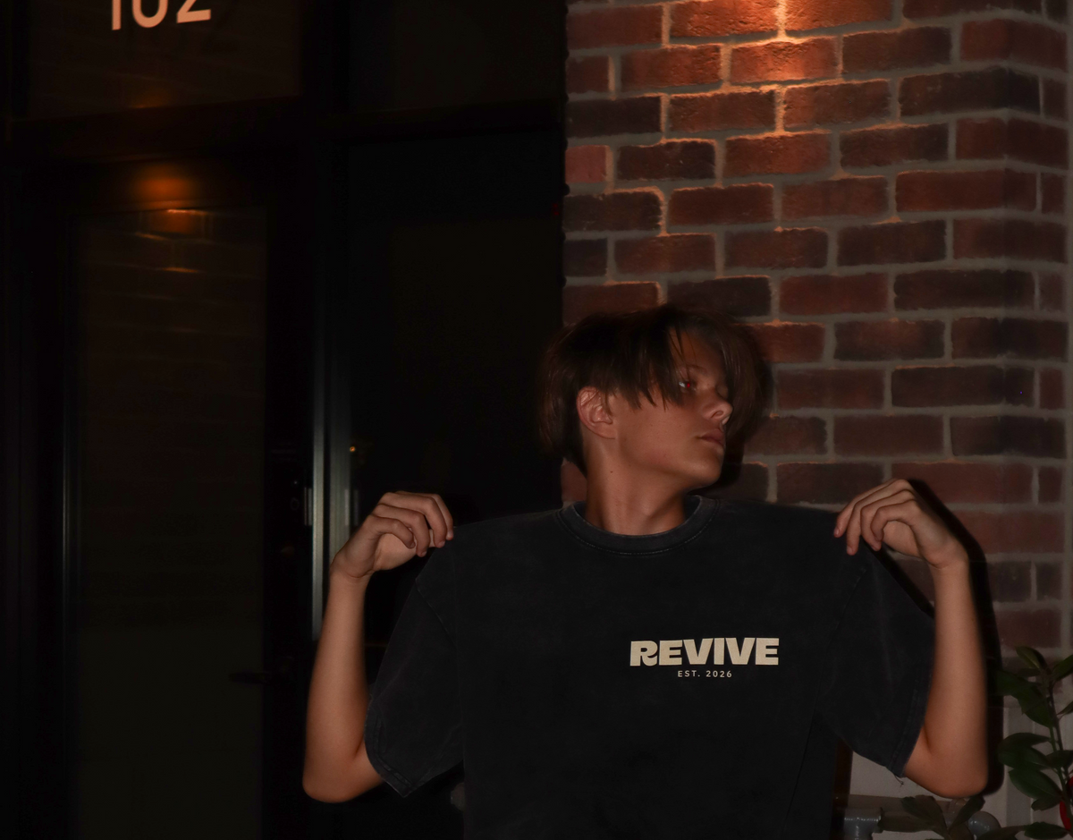 A person wearing a black t-shirt with 'REVIVE' printed in white on the chest, posing outdoors next to a red fire hydrant at night in front of a brick wall.
